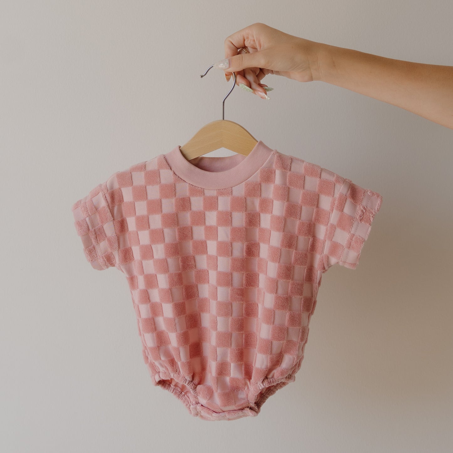 A hand displays the forever french baby Terry T-Shirt Romper in Sea Pink—a checkered, short-sleeved baby romper with easy snap closure—on a wooden hanger against a neutral background.