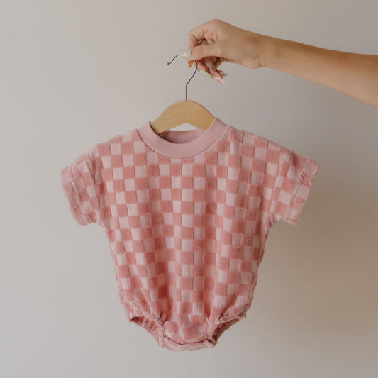 A hand displays the forever french baby Terry T-Shirt Romper in Sea Pink—a checkered, short-sleeved baby romper with easy snap closure—on a wooden hanger against a neutral background.