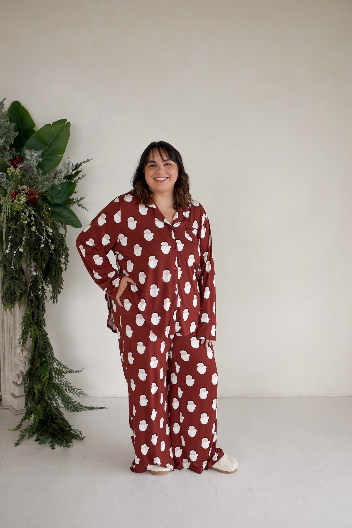 A woman smiles while wearing a red Women’s Bamboo Button Up Lounge Set | Vintage Santa, featuring a long-sleeved button-up shirt and matching wide-leg pants with a white vintage Santa print.