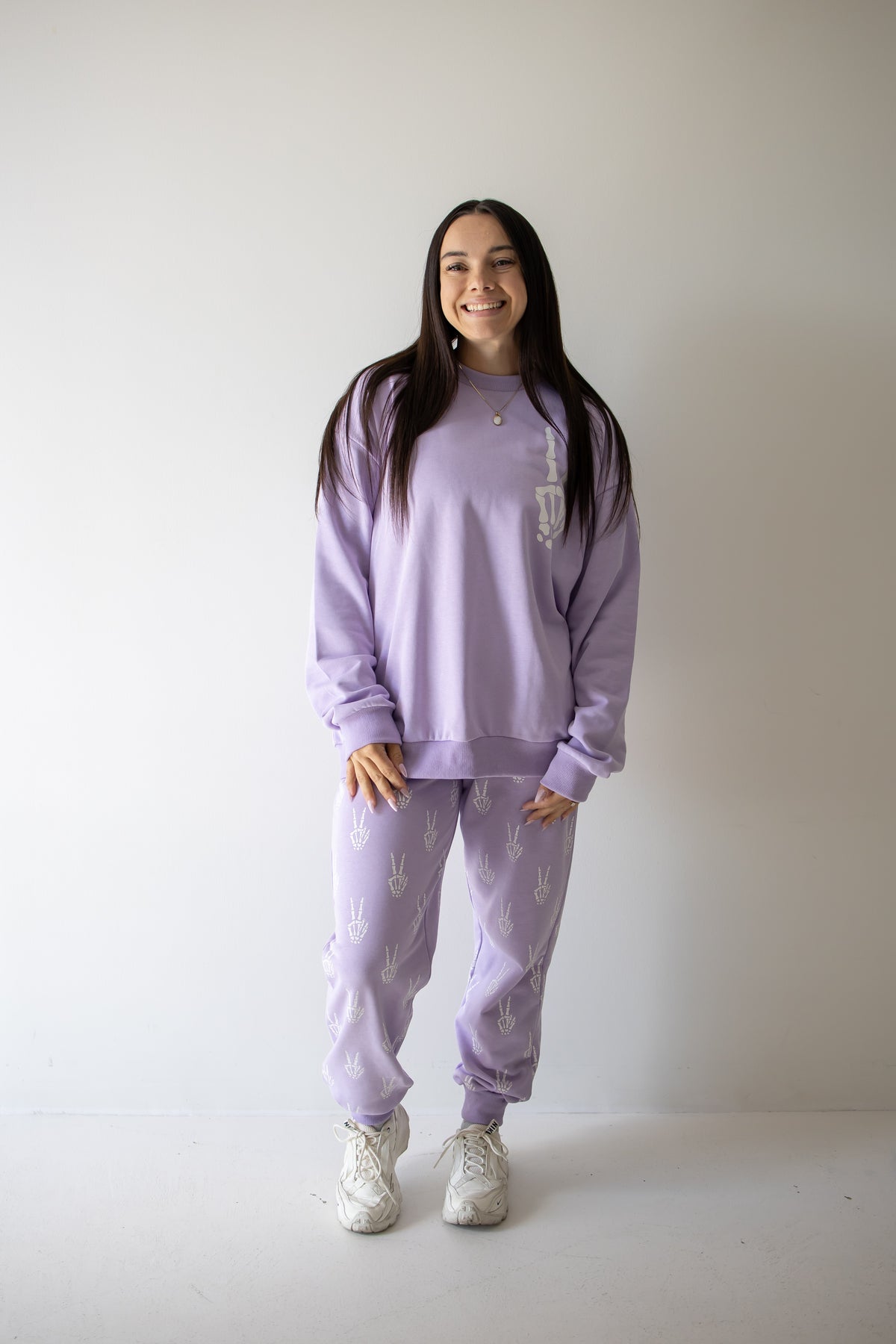 A woman stands against a plain white wall, smiling as she wears the forever french baby Women’s Sweat Set | Enchanted Skelly—a light purple ensemble adorned with whimsical white patterns and matching sweatpants. She pairs her cozy, stylish set with white sneakers, making it the perfect outfit for spooky season.
