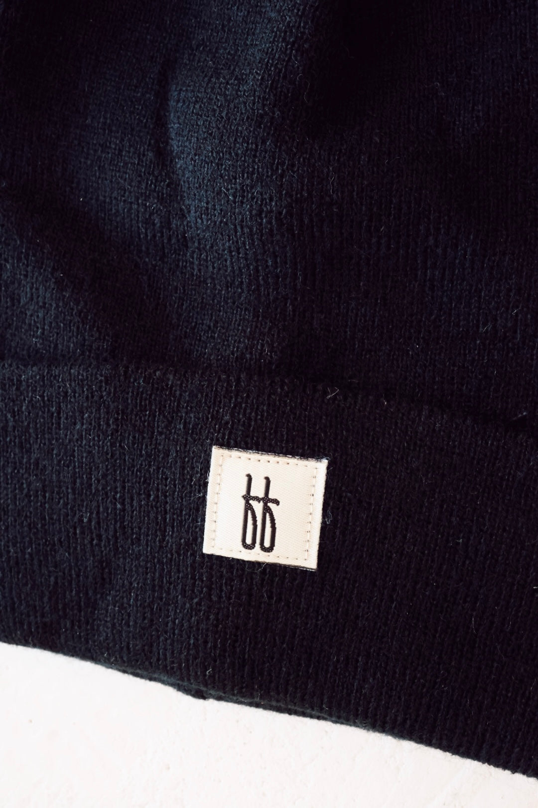 A close-up image of a black ribbed design, part of an "ff Knit Beanie | Adult" by forever french baby. The fabric features a small, rectangular white tag with a minimalist black logo in the center that resembles two stylized lowercase "b" letters placed side by side.