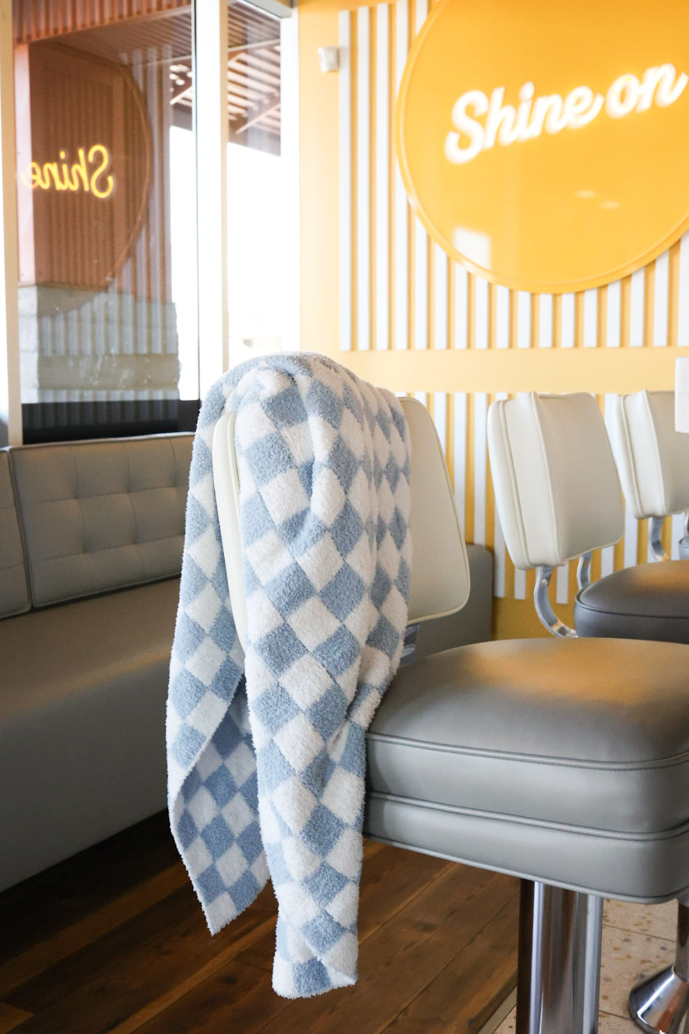 The forever french baby Plush Blanket | Morning Check, a soft blue-and-white checkered Oeko-Tex certified blanket, is draped over a modern gray bar stool in a bright room with light furniture and a yellow "Shine on" sign.