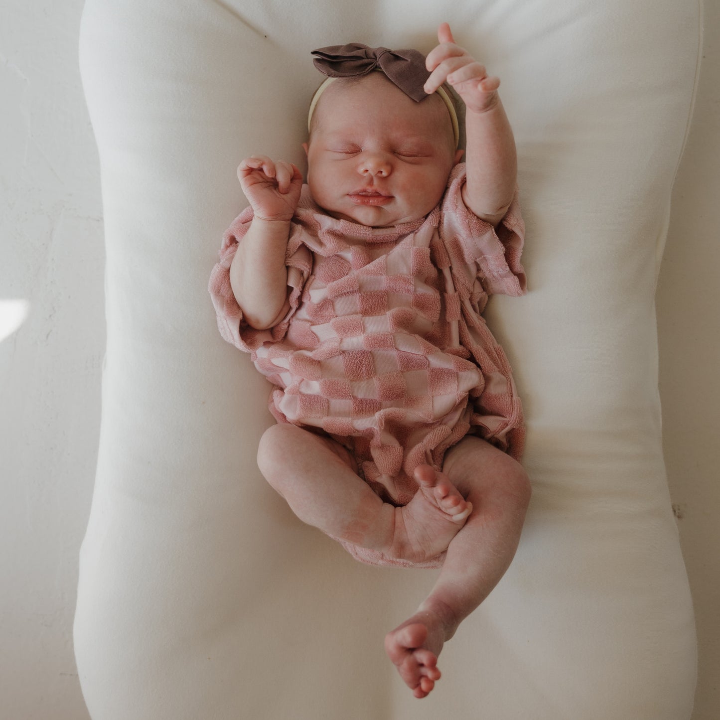 A newborn sleeps on a white cushioned surface in the forever french baby Terry T-Shirt Romper | Sea Pink, with snap closure and a brown bow headband—ideal for family matching outfits.
