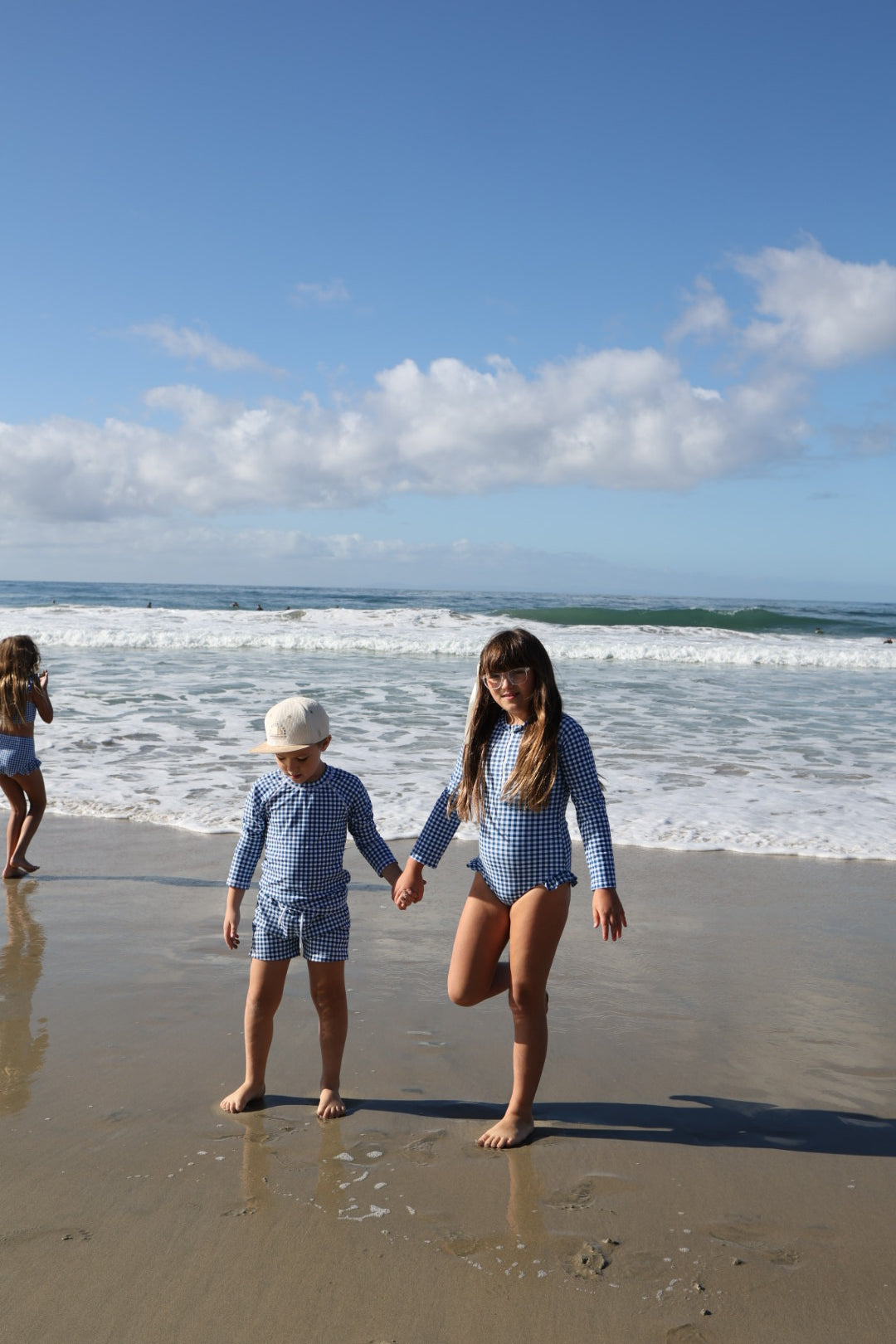 Two girls in forever french baby's blue gingham Girls Long Sleeve Swimsuit hold hands on a sandy beach, waves nearby. In the background, two more children play under a partly cloudy blue sky.