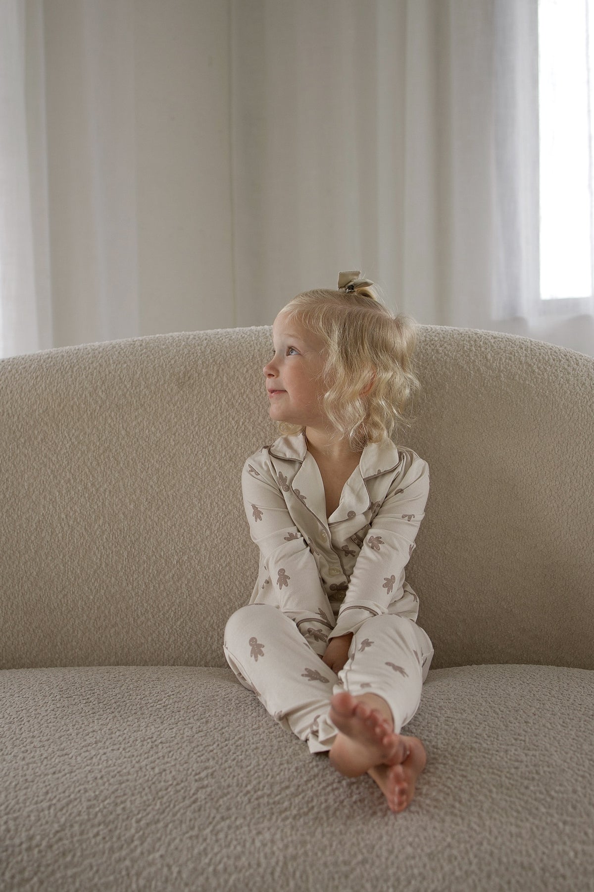 A young girl wearing a Girl’s Bamboo Button Up Lounge Set | Gingersnap with a brown gingerbread man print sits on a textured sofa.