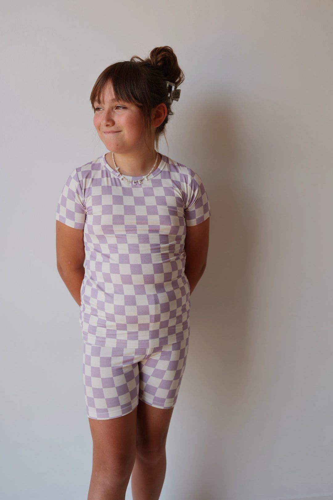 A young girl smiles in front of a light wall, wearing the forever french baby Bamboo Short Set | Grape Soda—a breathable, pastel purple and white checkerboard sleepwear perfect for cozy nights.