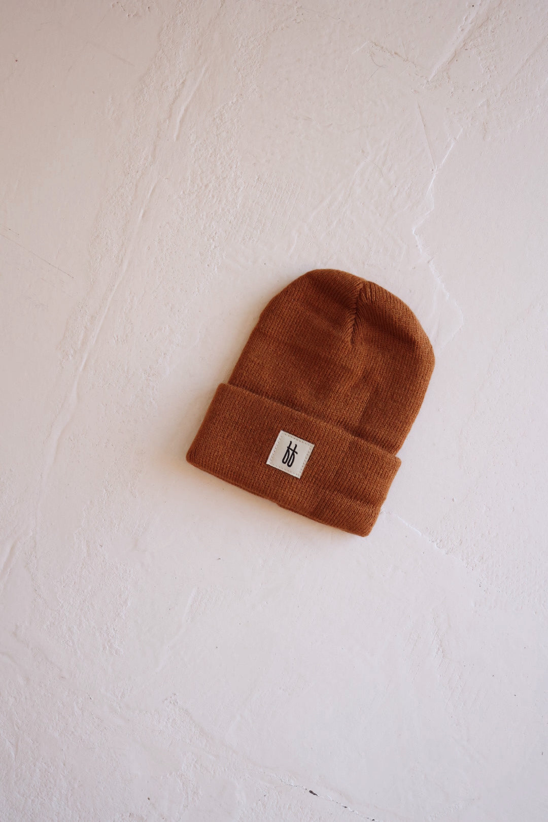 A rust-colored FF Knit Beanie from Forever French Baby, featuring a small white rectangular patch with a minimalist logo, is placed on a textured white surface. The beanie, true to Forever French Baby's design ethos, has a folded cuff and exhibits a simple, clean aesthetic.