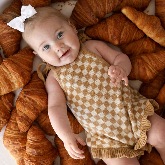 A baby with a white bow wears the forever french baby Knit Ruffle Romper in Latte Check, lying on her back among several croissants and smiling up.