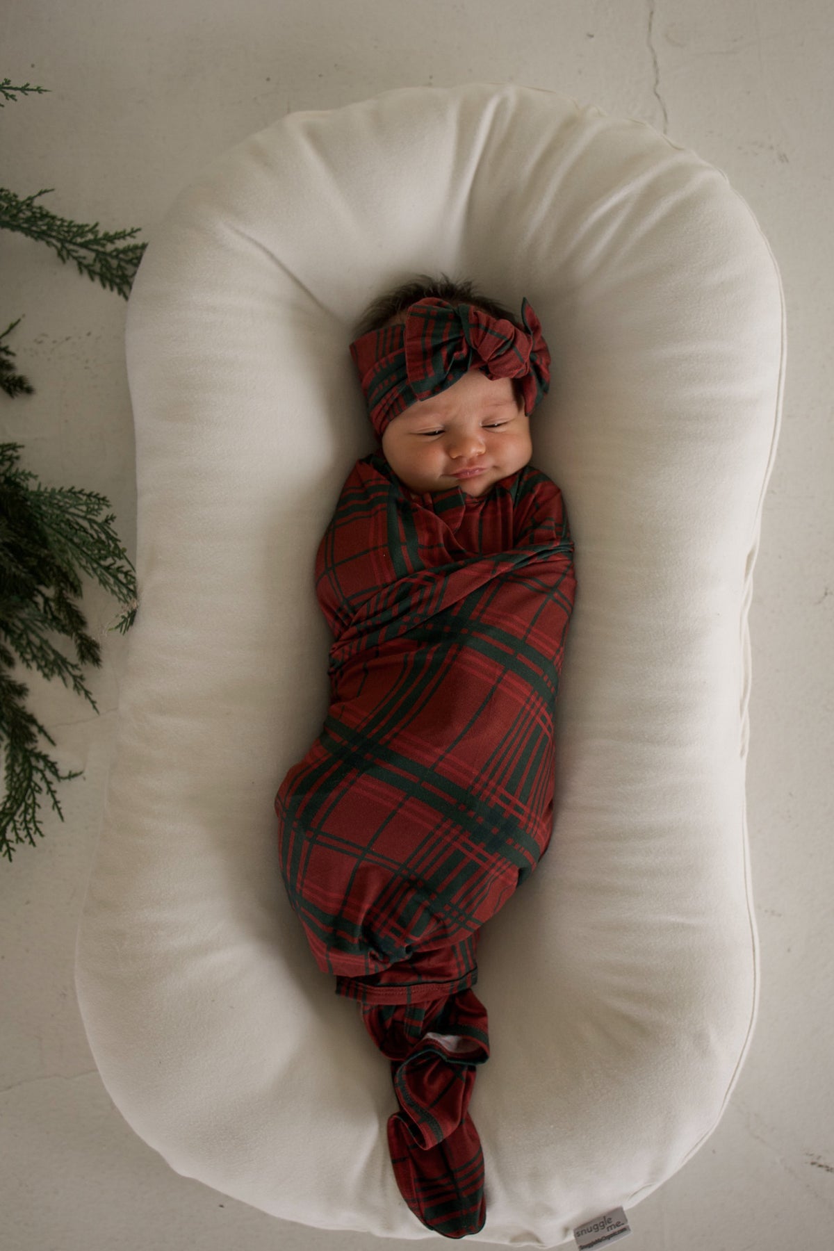 A newborn baby swaddled in a forever french baby Bamboo Swaddle | Christmas Plaid and matching knotted headband, resting in a white cushioned infant lounger.