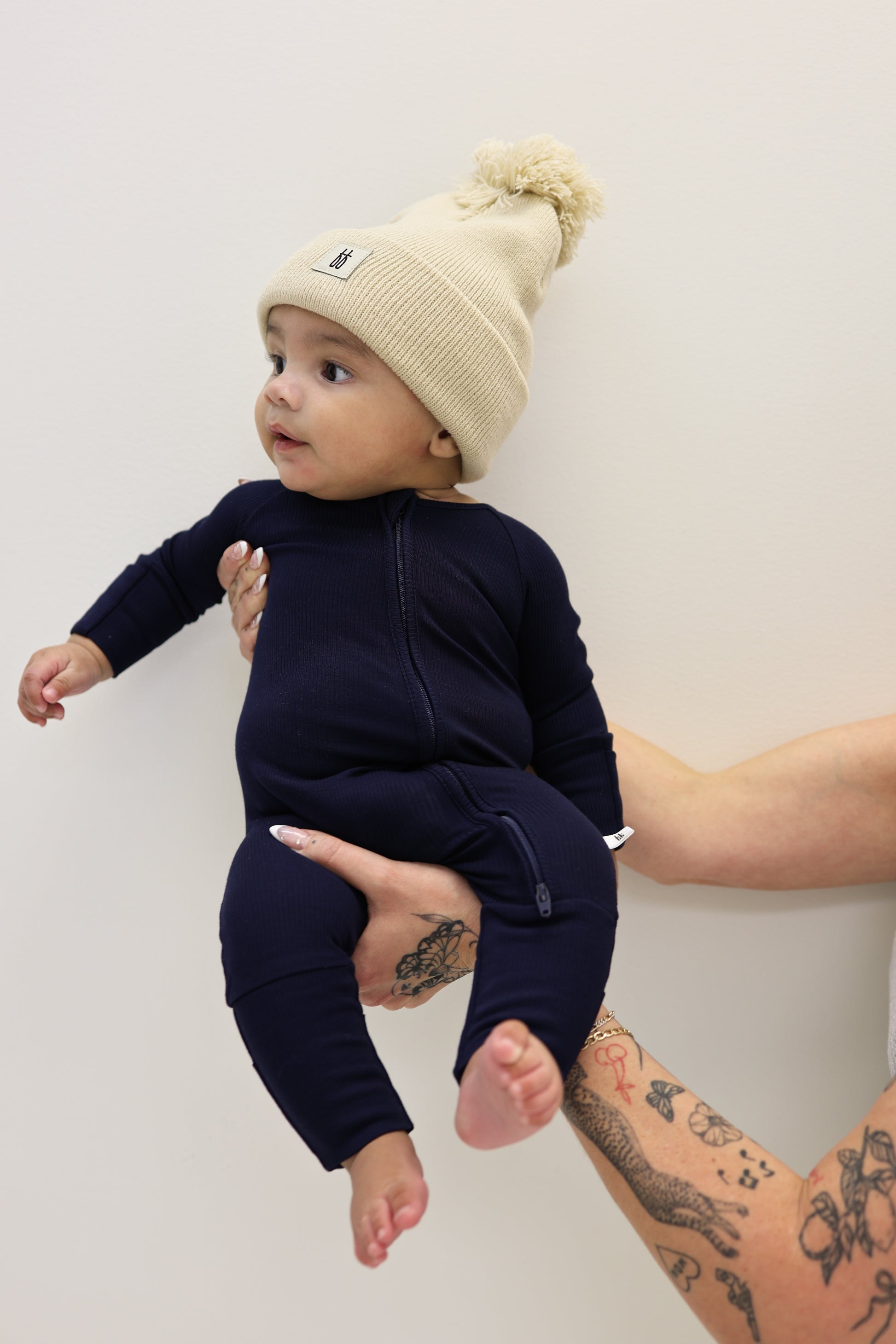 A baby in forever french baby Bamboo Zip Pajamas | Blueberry Ribbed and a cream knit beanie is held sideways by a tattooed adult, set against a plain light background.