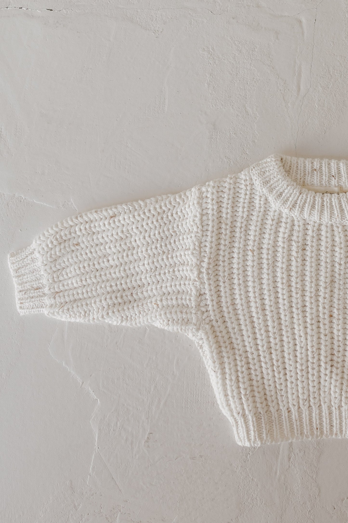 A close-up of the forever french baby Child Knit Sweater | Cloud Confetti, cream-colored with a ribbed texture, is laid flat on a light surface, showing only the upper body and one long sleeve.
