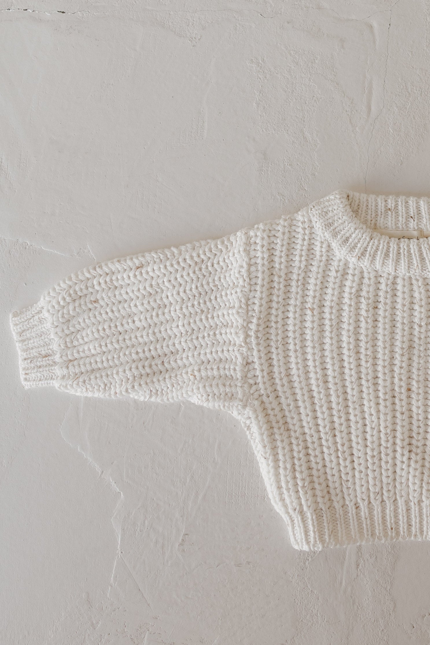 A close-up of the forever french baby Child Knit Sweater | Cloud Confetti, cream-colored with a ribbed texture, is laid flat on a light surface, showing only the upper body and one long sleeve.