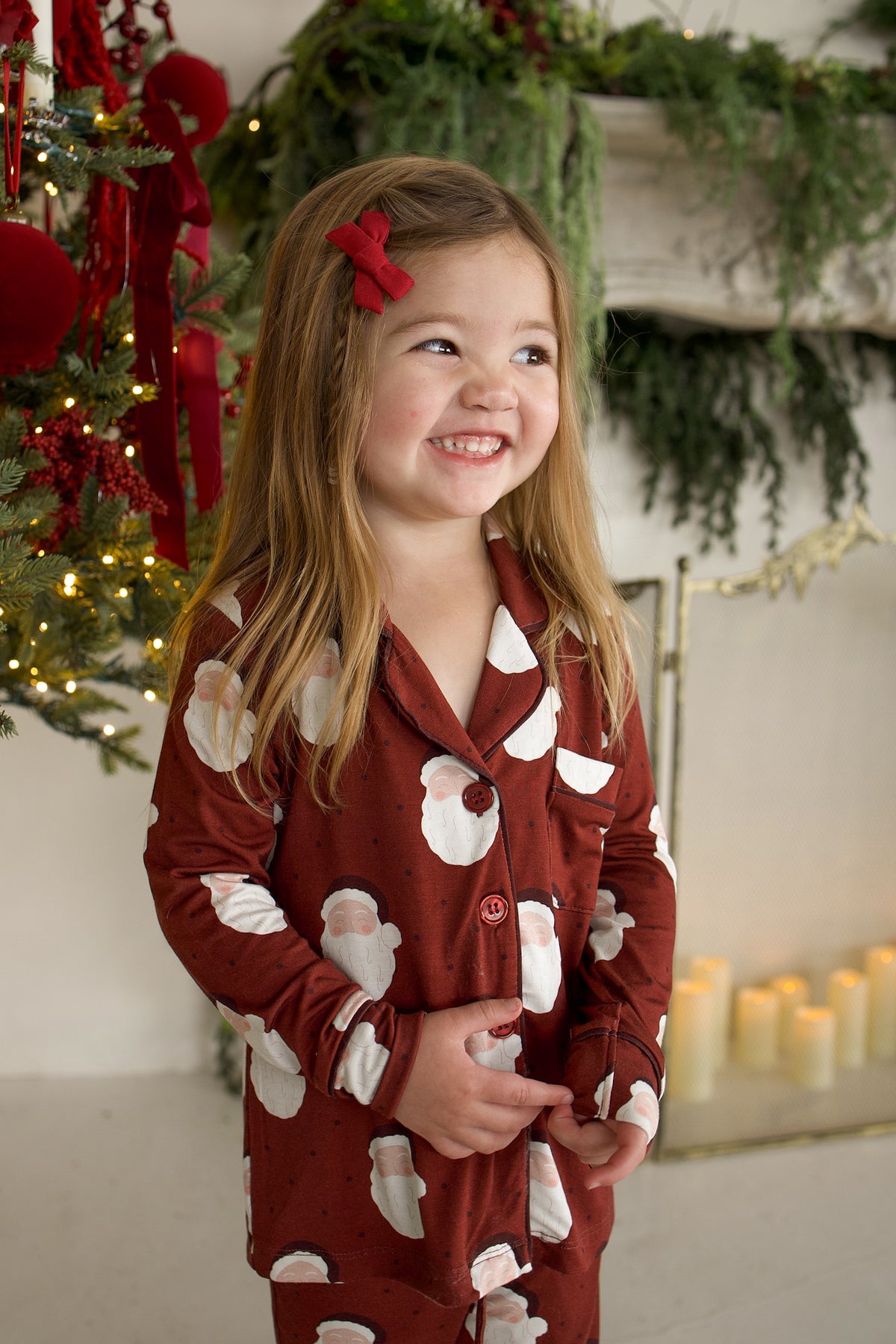 A young girl smiling while wearing a forever french baby Girl’s Bamboo Button Up Lounge Set in Vintage Santa, featuring a long-sleeved top and pants with a vintage Santa Claus print.
