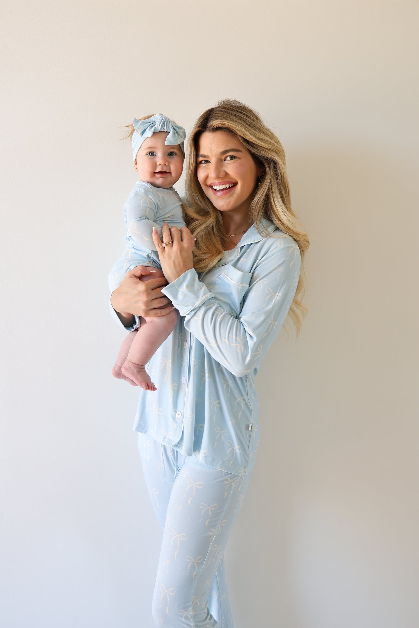 A woman in a light blue forever french baby Women's Bamboo Button Up Lounge Set | Blue Bows holds a smiling baby in matching blue pajamas and headband, both standing against a plain light background.