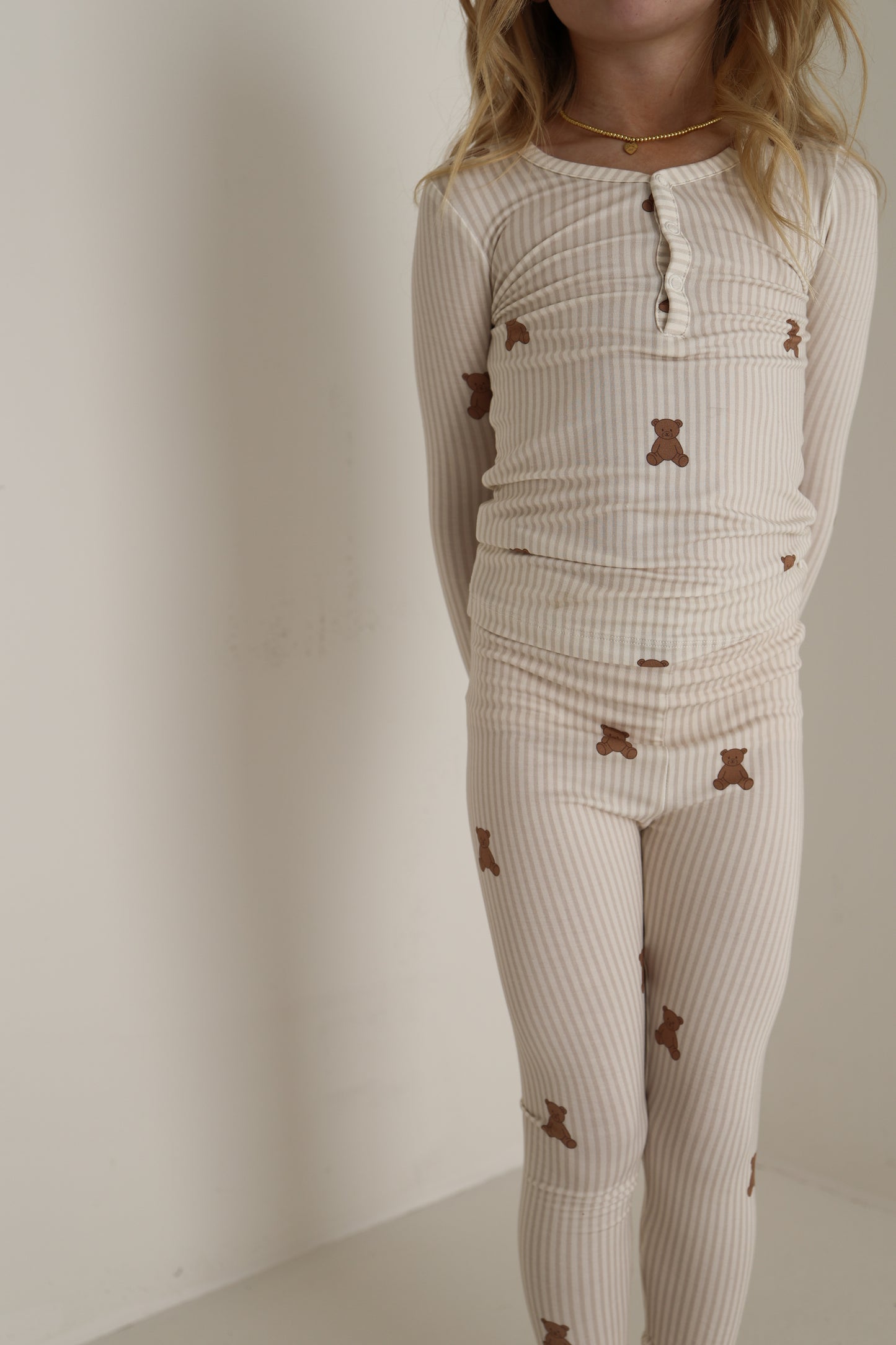 A child stands against a plain, light wall wearing forever french baby's Bamboo Two Piece Pajamas | Teddy, which are cream-colored with ribbed fabric and small brown teddy bear prints. The child's face is not visible.