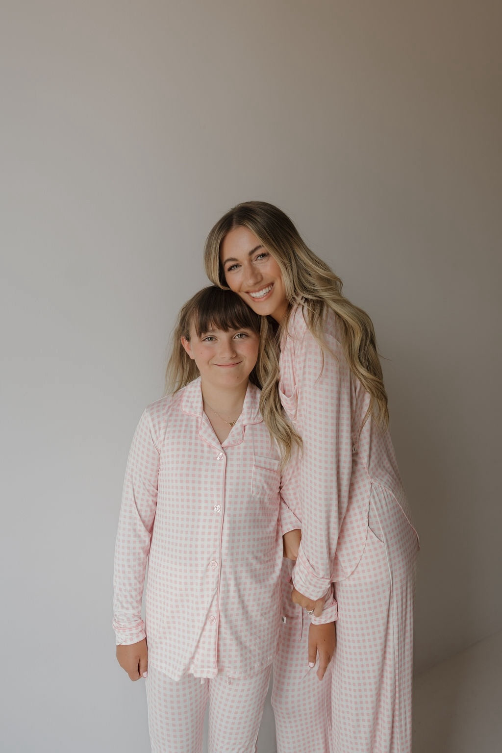 A smiling woman and a young girl, both dressed in matching pink and white checkered pajamas, stand close together against a plain light background. The woman is wearing the forever french baby Women’s Bamboo Button Up Lounge Set | Giselle, which features wide leg lounge pants for a cozy yet stylish coordinated look.