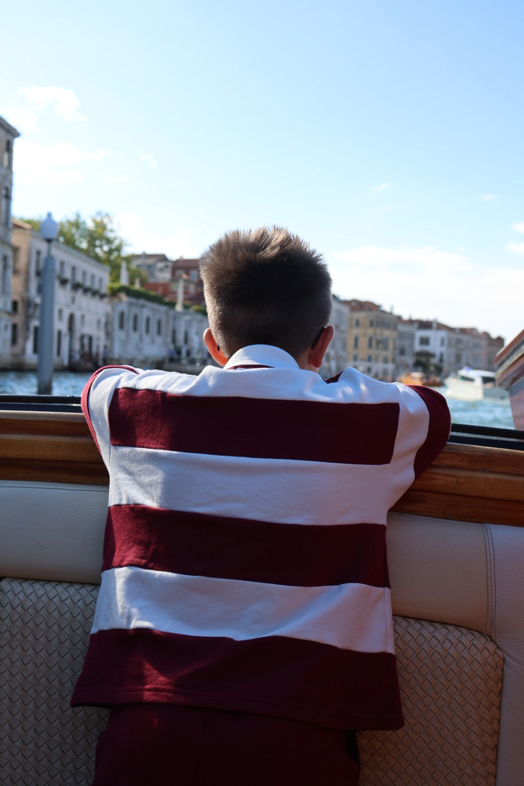 A child in a burgundy and white Child Rugby Sweatshirt | Deep Cranberry looking out from a boat at buildings along a canal.