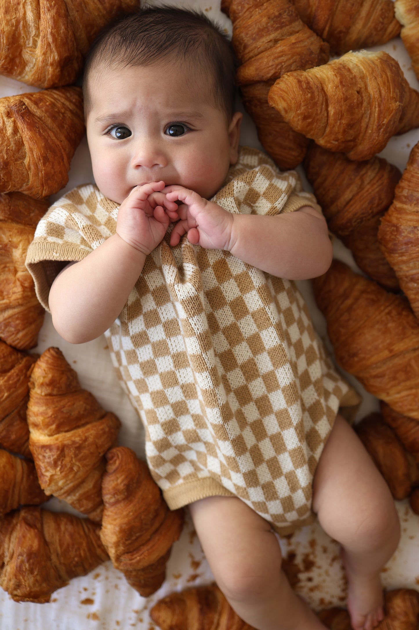 A baby wearing the Knit Romper | Latte Check by forever french baby lies on a white surface, surrounded by croissants, looking up with a slightly worried expression and hands near their mouth.