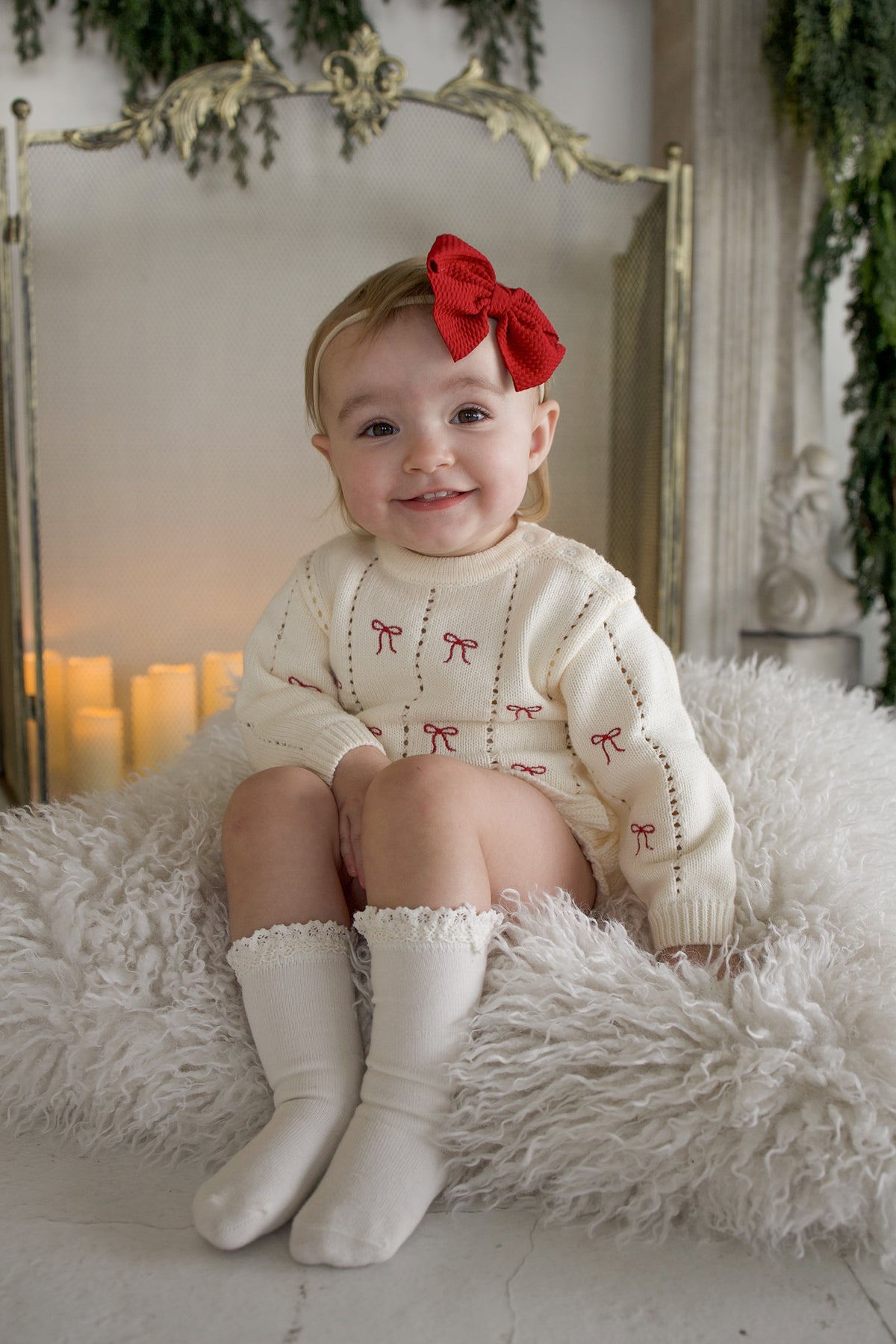 A smiling toddler wearing a Knit Ruffle Romper | Candy Cane with knee-high socks and a large red hair bow.