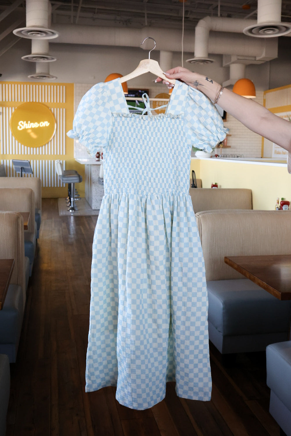 The Women's Short Sleeve Muslin Dress in Morning Check by forever french baby, featuring light blue and white checks and puff sleeves, hangs on a hanger in a retro diner with booth seating and wooden floors.