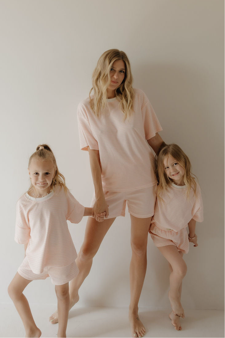 A woman and two girls stand barefoot against a plain light wall, holding hands and smiling as they wear matching forever french baby Women's Terry Short Set in Cotton Candy.