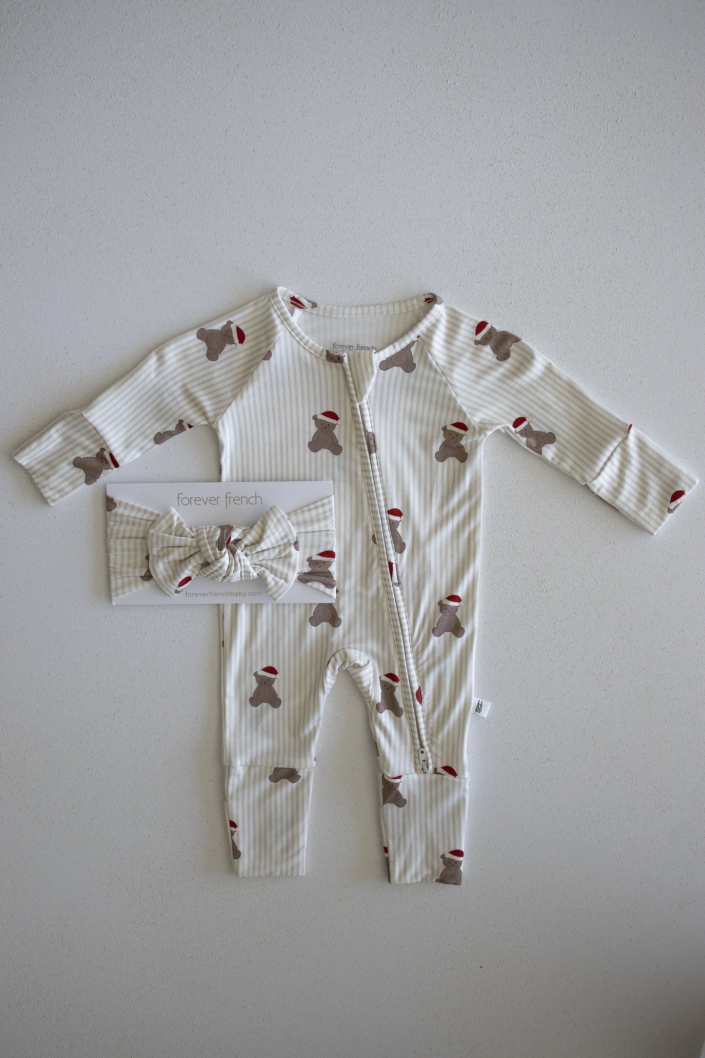 Introducing the Bamboo Zip Pajamas | Santa Teddy by forever french baby: a cozy baby outfit featuring beige and white stripes with charming brown teddy bear and festive red Santa hat print, laid flat. Crafted from hypoallergenic materials gentle for delicate skin. A matching bow headband is packaged to the left of the romper for a coordinated look.