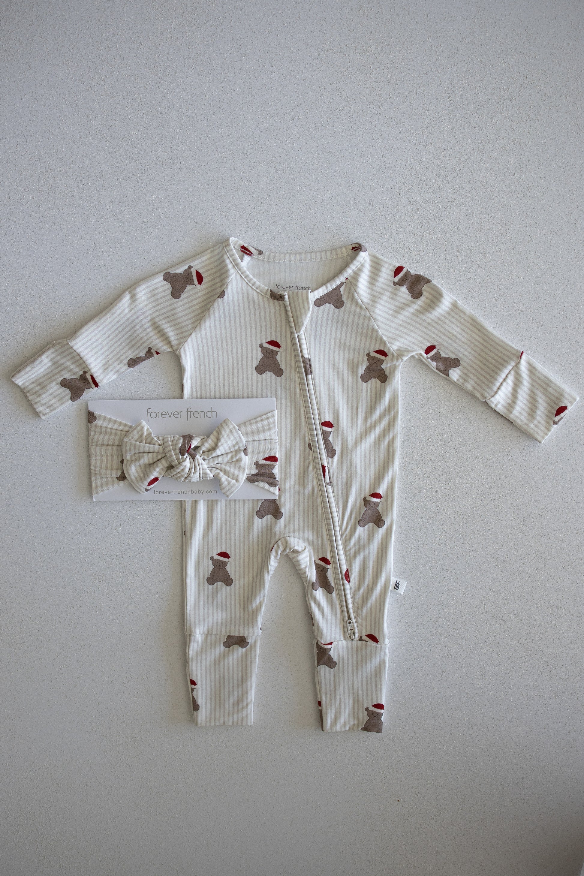 Introducing the Bamboo Zip Pajamas | Santa Teddy by forever french baby: a cozy baby outfit featuring beige and white stripes with charming brown teddy bear and festive red Santa hat print, laid flat. Crafted from hypoallergenic materials gentle for delicate skin. A matching bow headband is packaged to the left of the romper for a coordinated look.