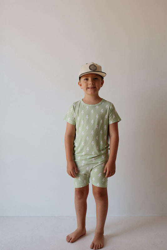 A young child stands barefoot against a light wall, wearing the forever french baby Bamboo Short Set | Flash—light green, hypoallergenic matching tee and shorts with white lightning bolts—and a beige baseball cap.