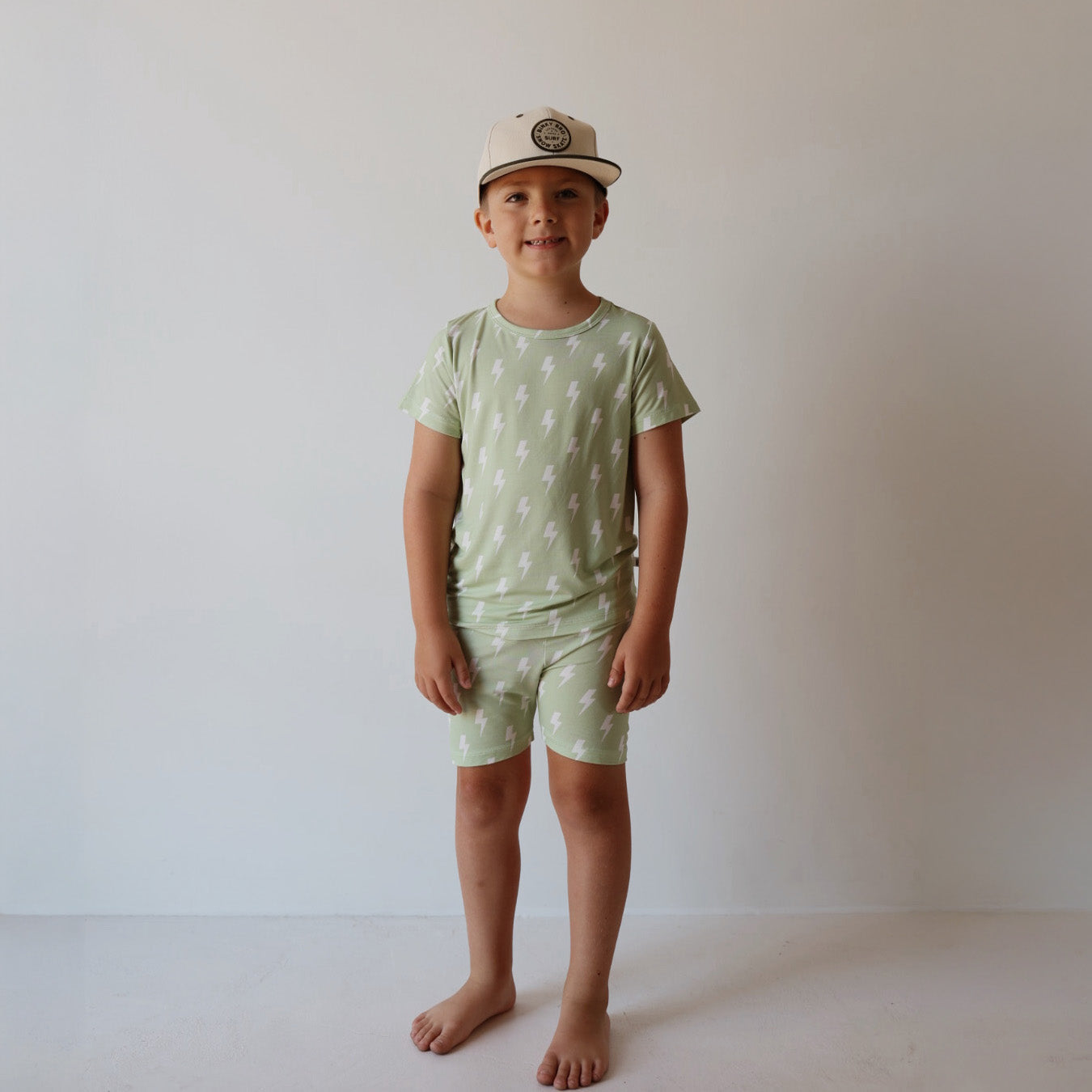 A young child stands barefoot against a white background, wearing the forever french baby Bamboo Short Set | Flash—light green matching t-shirt and shorts with white lightning bolts—hypo-allergenic, stylish, and comfy, plus a light baseball cap.
