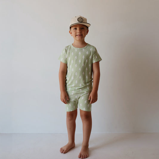 A young child stands barefoot against a white background, wearing the forever french baby Bamboo Short Set | Flash—light green matching t-shirt and shorts with white lightning bolts—hypo-allergenic, stylish, and comfy, plus a light baseball cap.