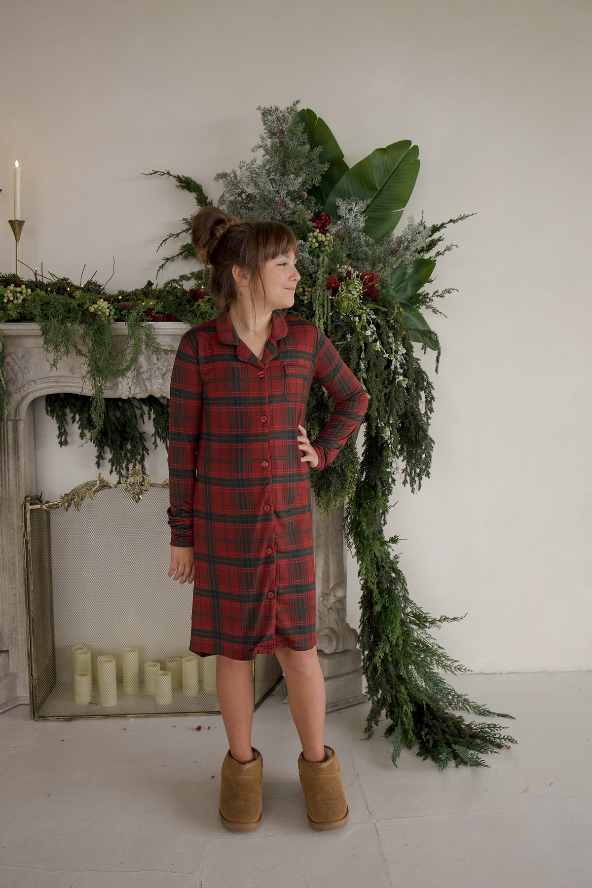 A girl wears a Bamboo Sleeping Dress | Christmas Plaid button-down nightshirt and tan boots while standing in front of a fireplace.