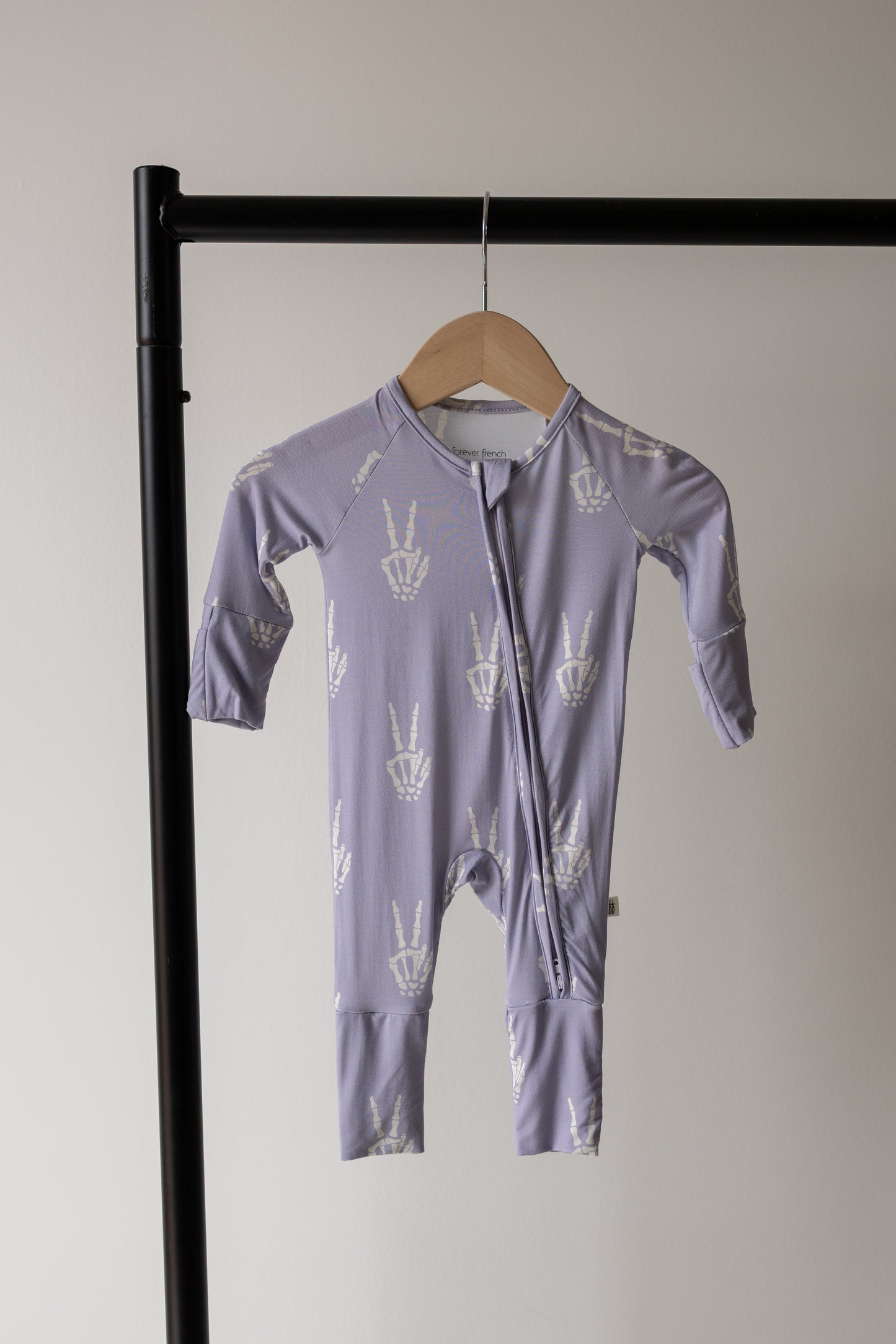 The forever french baby Bamboo Zip Pajamas | Enchanted Skelly, featuring a lavender design with white peace sign prints and crafted from ultra-soft hypo-allergenic bamboo viscose, hangs on a wooden hanger against a plain light wall and black clothing rack.