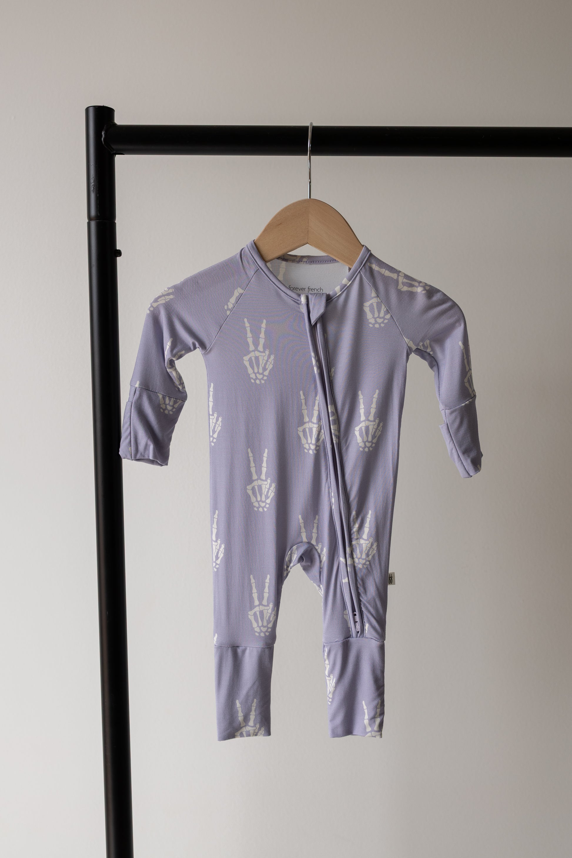 The forever french baby Bamboo Zip Pajamas | Enchanted Skelly, featuring a lavender design with white peace sign prints and crafted from ultra-soft hypo-allergenic bamboo viscose, hangs on a wooden hanger against a plain light wall and black clothing rack.