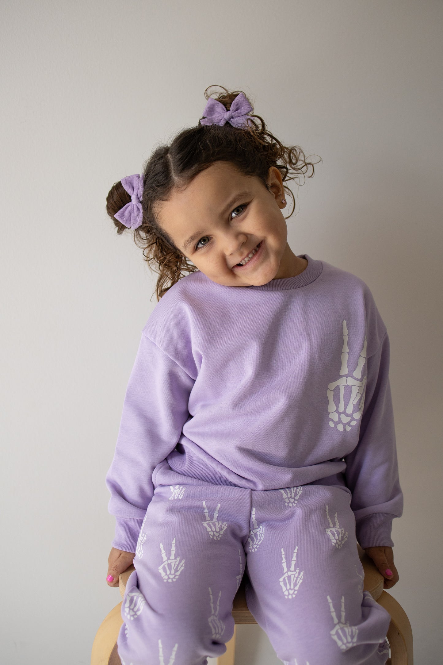 A young girl with curly hair in purple bows smiles, wearing the forever french baby Child Sweat Set | Enchanted Skelly—a matching lavender outfit featuring white skeleton peace sign prints—as she sits on a wooden stool against a plain light background, ready for everyday adventures.