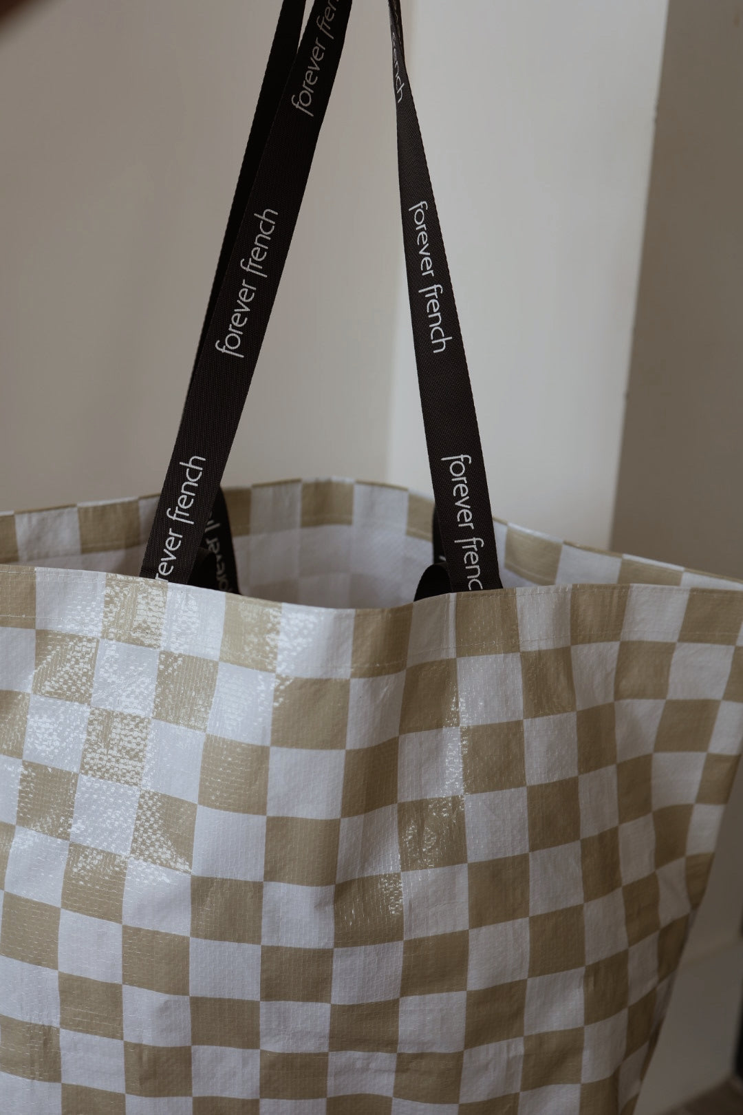 The ff Warehouse Sale Tote by forever french baby features a large beige and white checkered design, shiny woven material, and black handles printed with "forever french" in white. The bag is displayed against a plain wall.