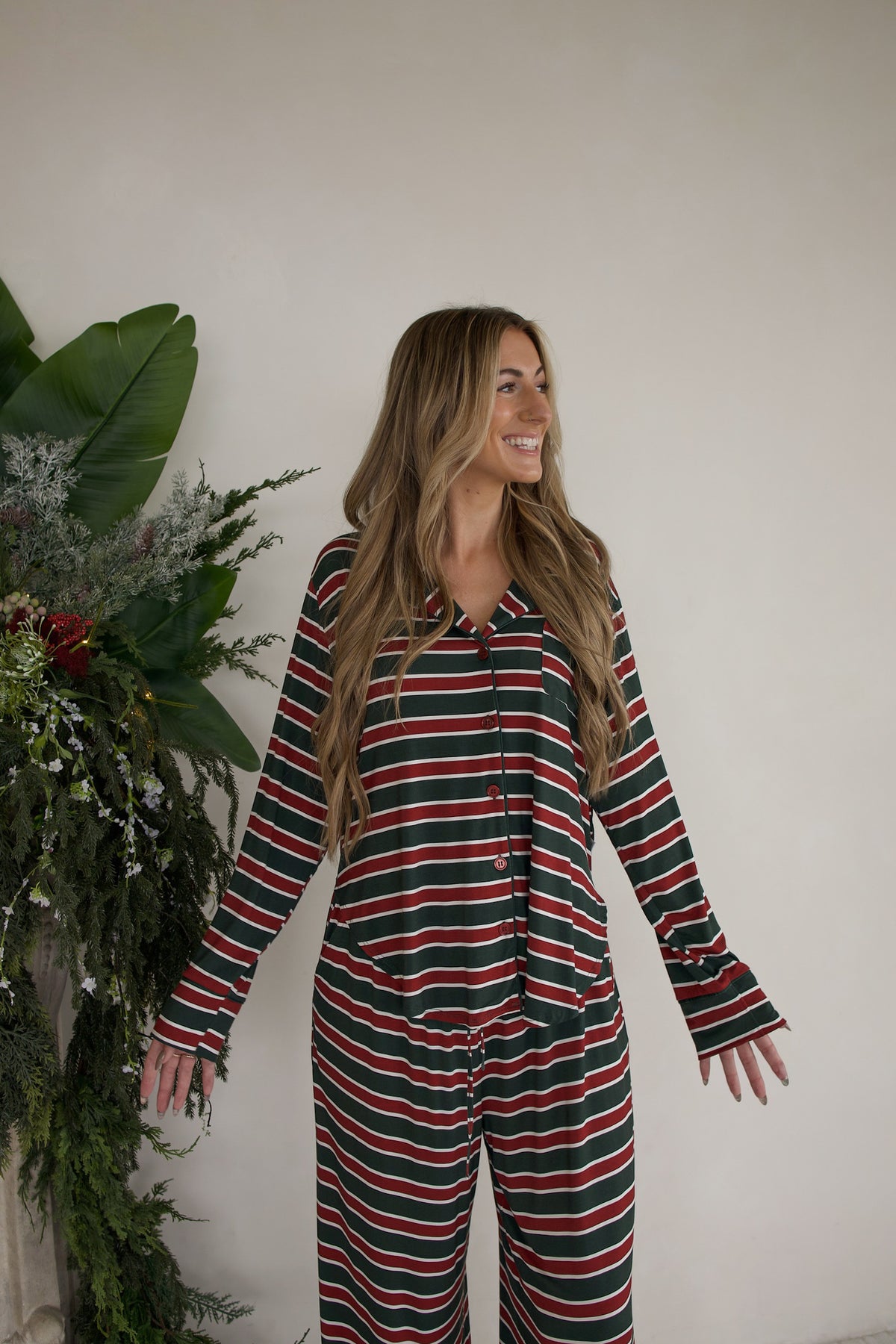 A smiling woman wearing a Women’s Bamboo Button Up Lounge Set | Holiday Stripe featuring a long-sleeved button-up top and matching wide leg lounge pants in horizontal red, green, and white stripes.