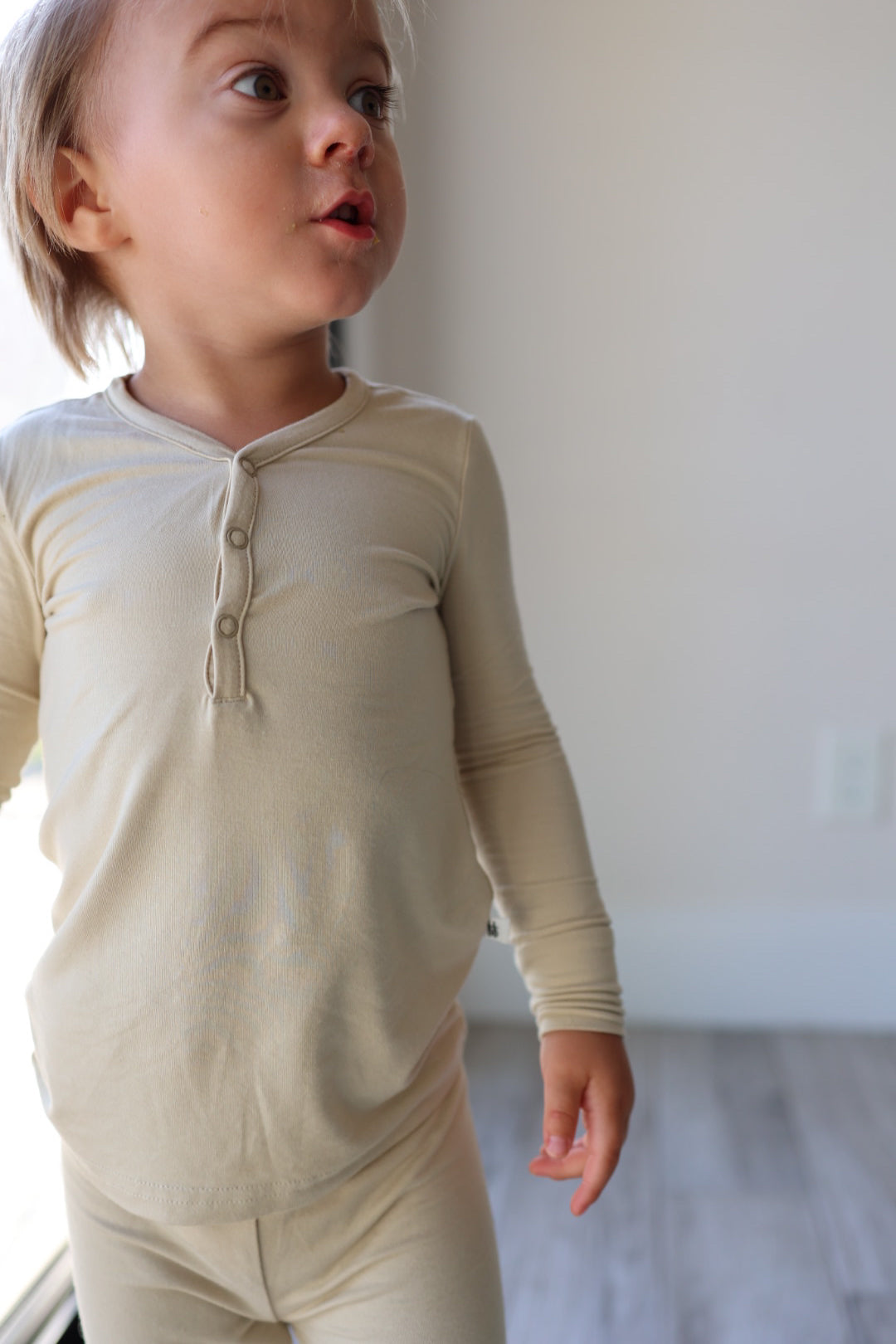 A young child wearing Limestone-colored Bamboo Two Piece Pajamas from forever french baby stands near a window, looking to the side. The child has straight light brown hair and appears to be in a bright room with light-colored wooden flooring, showcasing the breathable, hypo-allergenic fabric of their clothing.