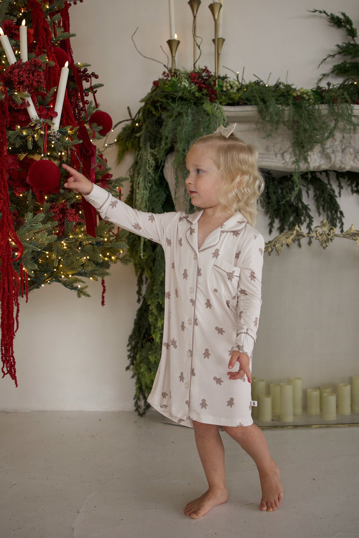 A toddler in a Bamboo Sleeping Dress | Gingersnap with a gingerbread man print points at an ornament on a Christmas tree.