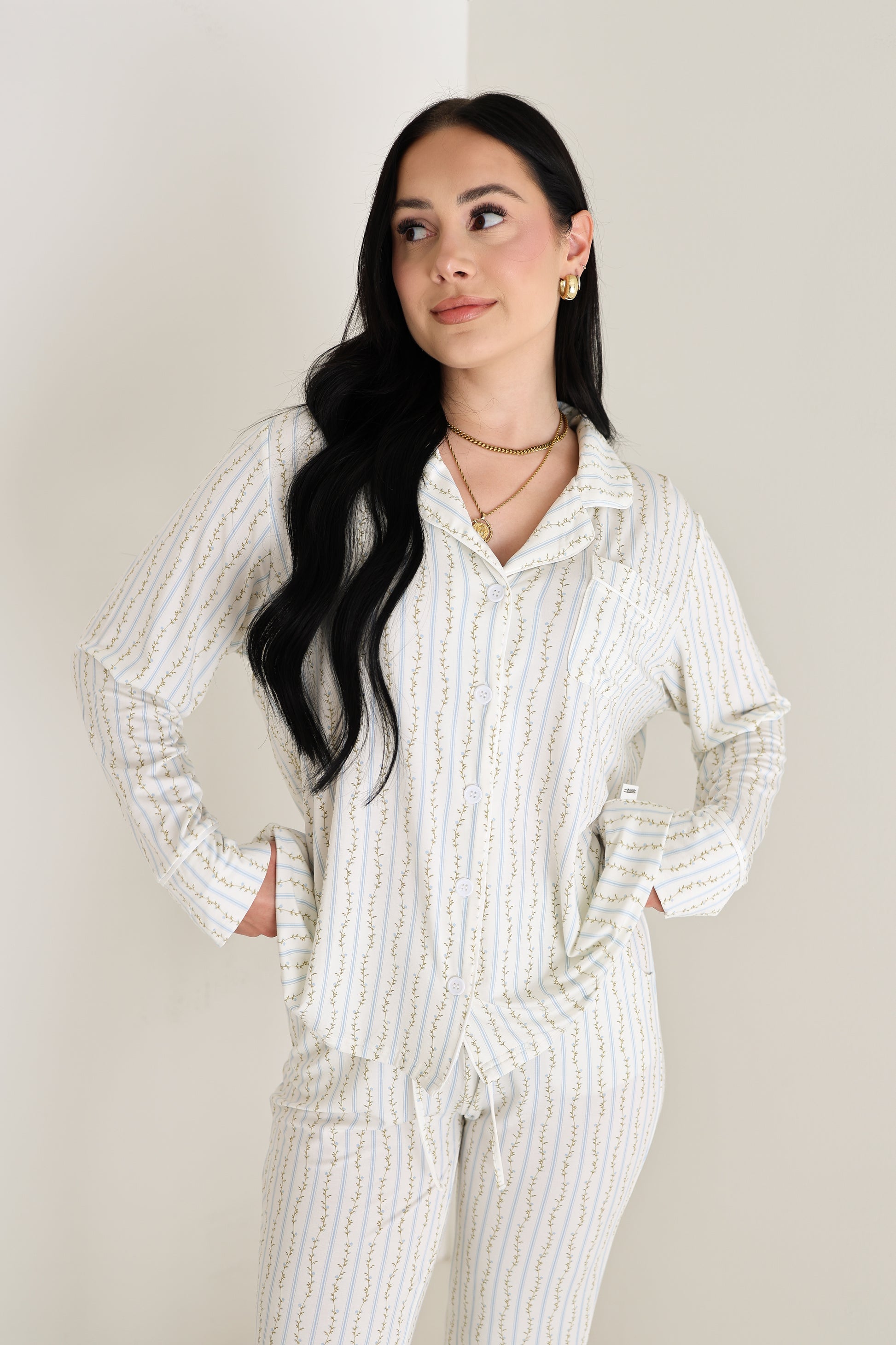 A woman with long black hair models the forever french baby Women's Bamboo Button Up Lounge Set | Dainty Vines, standing by a light wall with her hands in her pockets, gazing to the side and smiling in stylish, hypo-allergenic bamboo clothing.