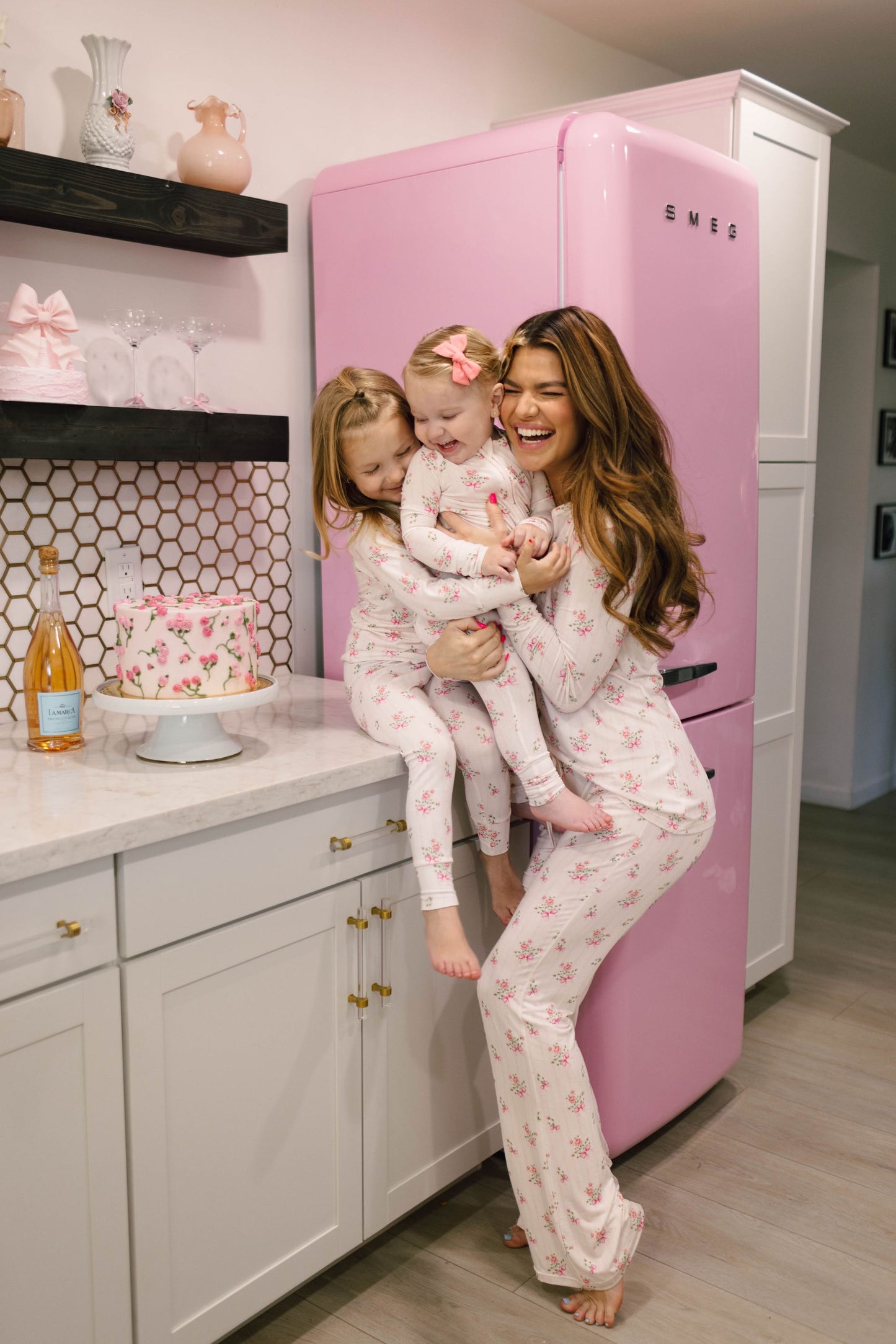 A woman and two young children share a joyful moment in the kitchen, all dressed in matching Women's Bamboo Pajamas from the Kendy Floral Collection by Kendy 2. The woman holds one child while the other stands nearby. A pink fridge, cake, and champagne bottle add to the cheerful and warm scene.