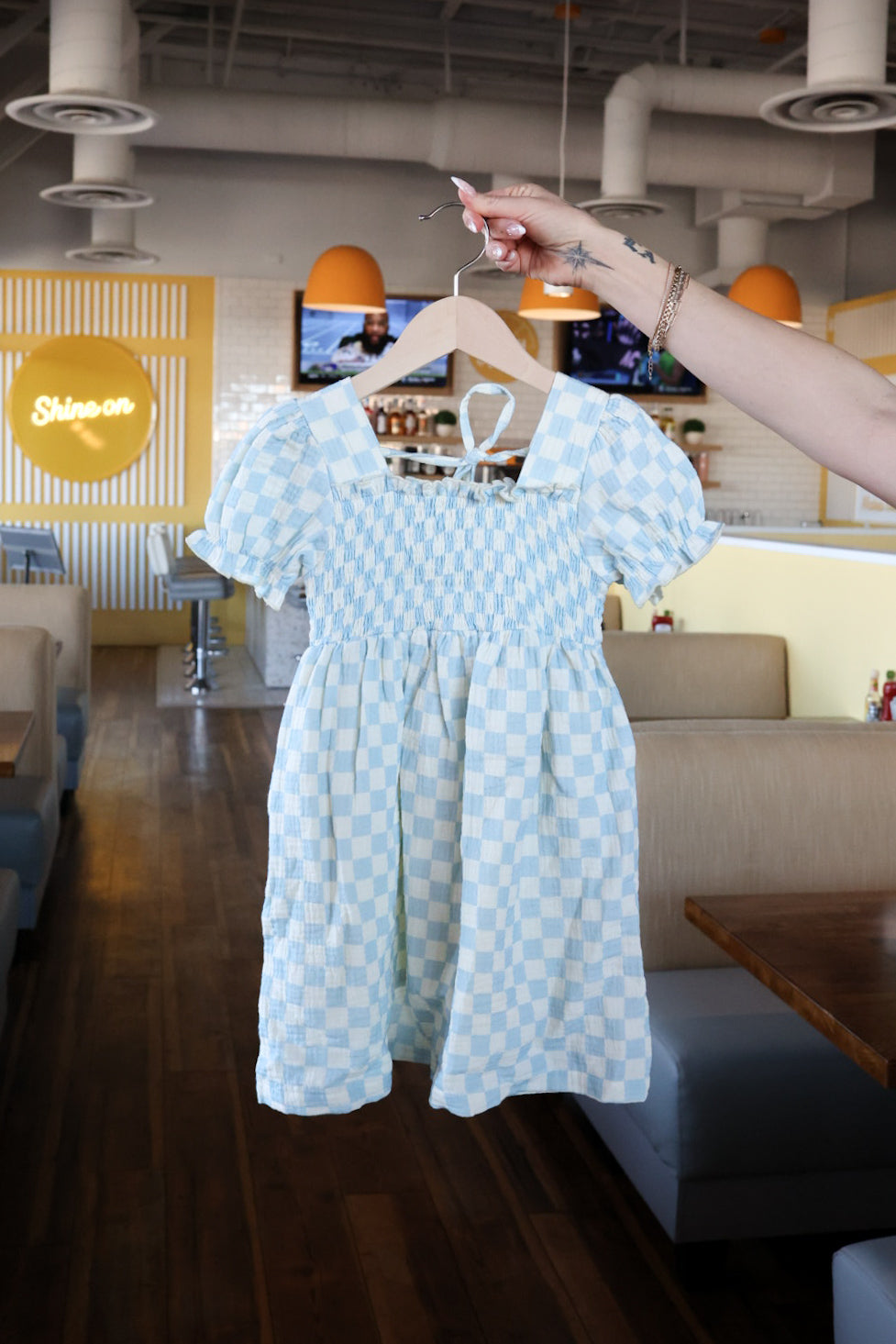 A hand holds a forever french baby Child Muslin Dress | Morning Check, featuring blue and white checks, inside a modern diner with wooden floors, booths, and a yellow "Shinon" sign.