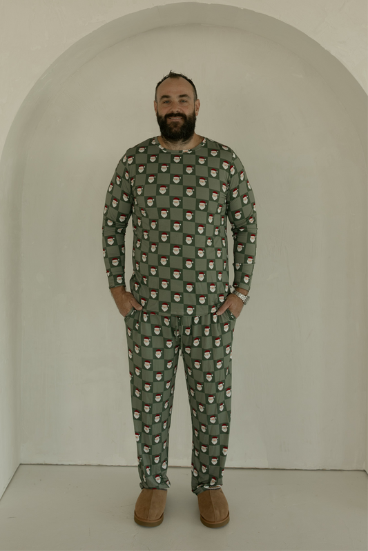 A bearded man smiles in a minimal white alcove, hands in his pockets, wearing forever french baby's Men's Bamboo Pajamas | Santa Checkerboard—festive green sleepwear that’s breathable and comfortable—with cozy brown slippers.