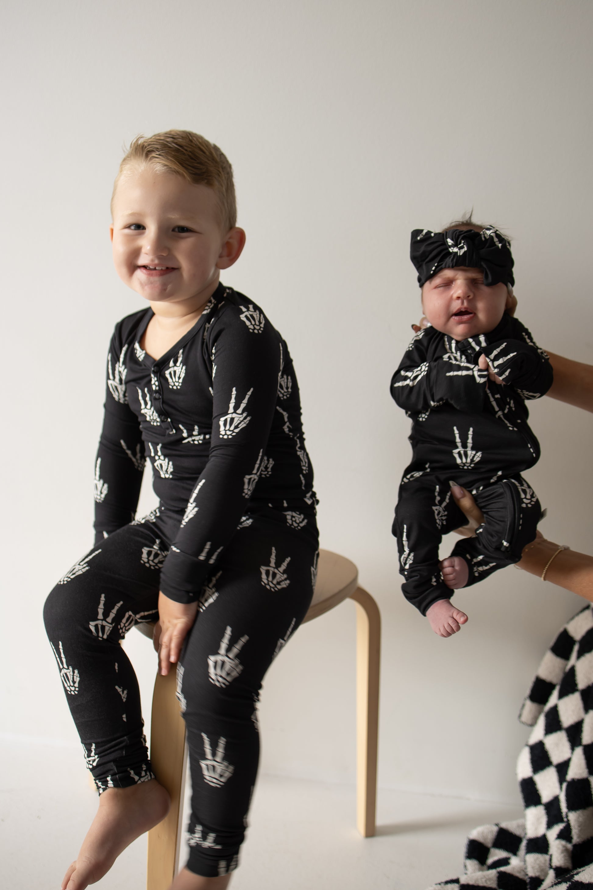 A young child sits on a wooden stool, smiling, next to a baby being held by an adult. Both children wear matching Midnight Skelly Bamboo Two Piece Pajamas from forever french baby, featuring white hand sign patterns and crafted from breathable bamboo fabric to keep little ones comfortable all day long.
