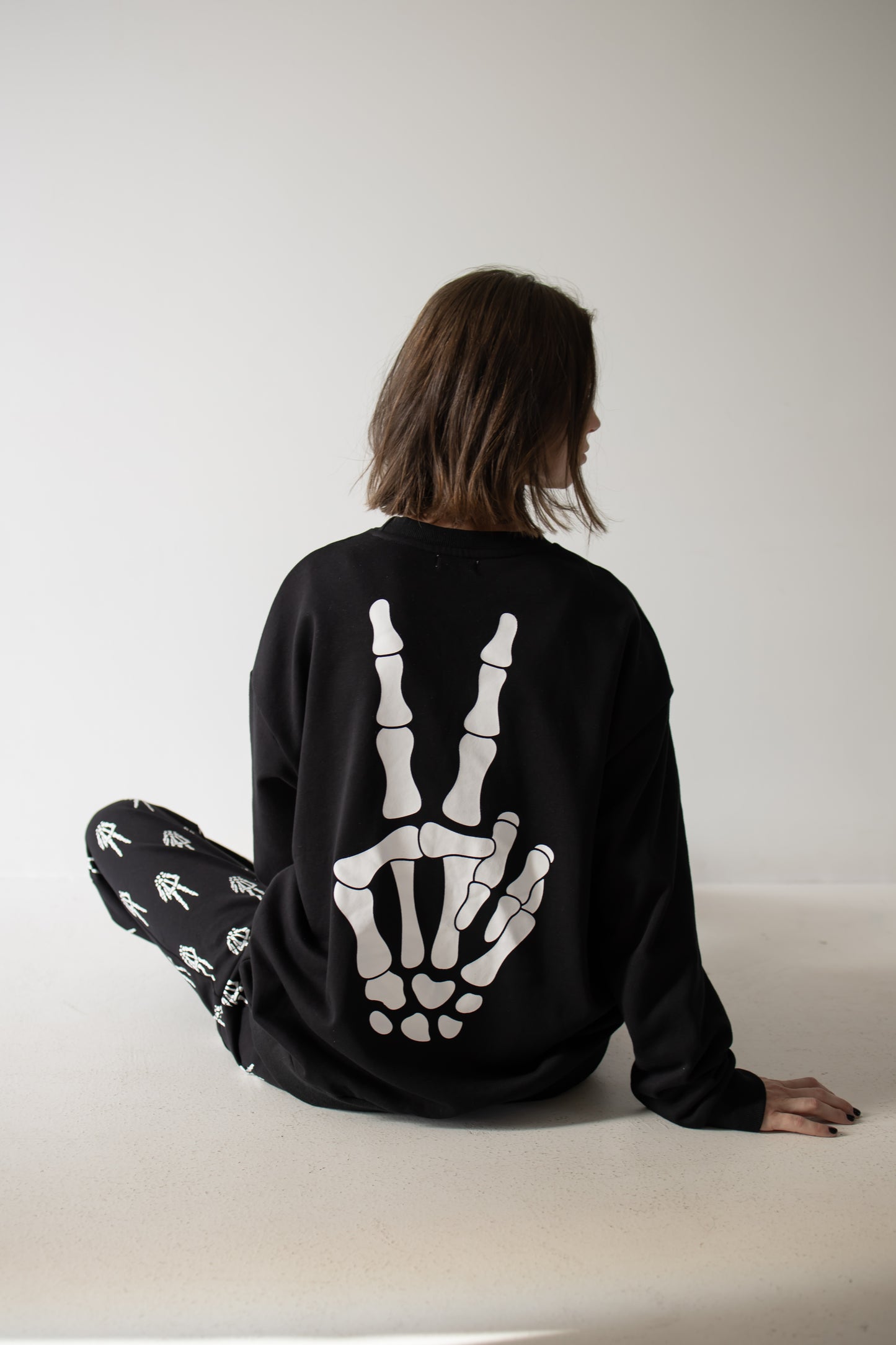 A person with brown hair sits on the floor, facing away. They wear the Women’s Sweat Set | Midnight Skelly by forever french baby—a black sweat set featuring a skeletal hand peace sign and matching black pants with white bone designs—making it an ideal pumpkin patch outfit for spooky season.