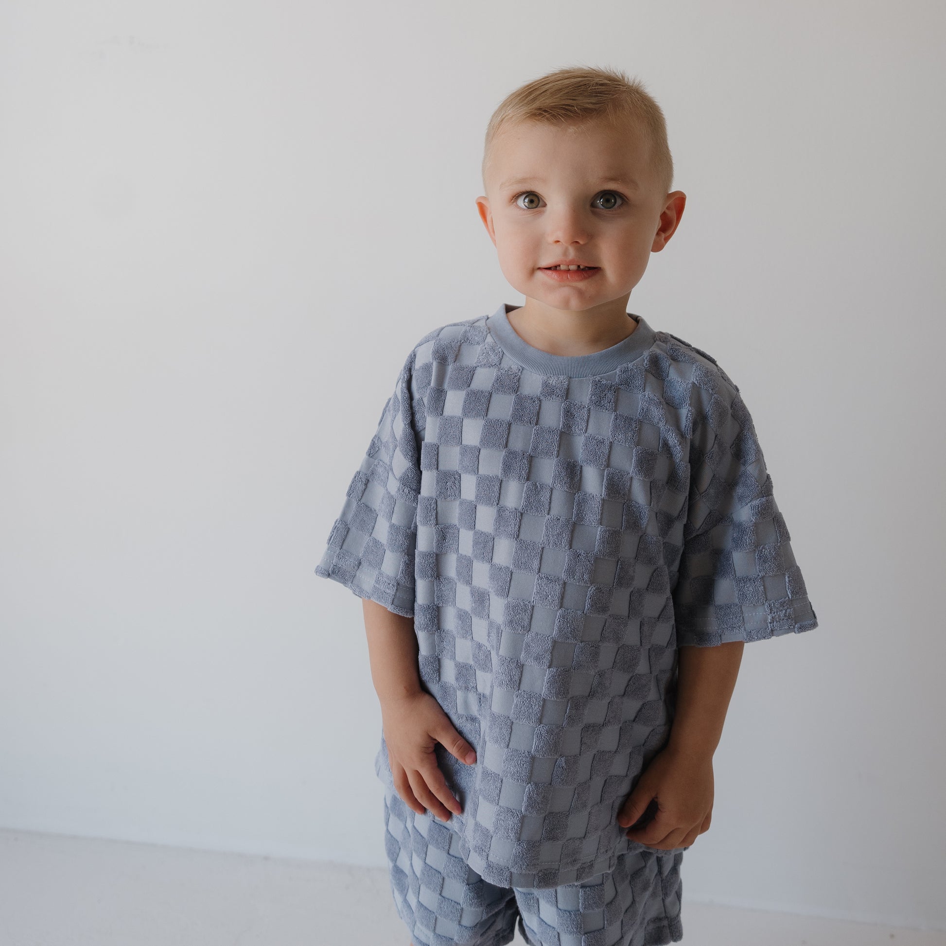 A young child with short blond hair wears the forever french baby Child Terry Short Set | Tradewinds, standing against a plain white background and looking slightly to the side with a neutral expression.