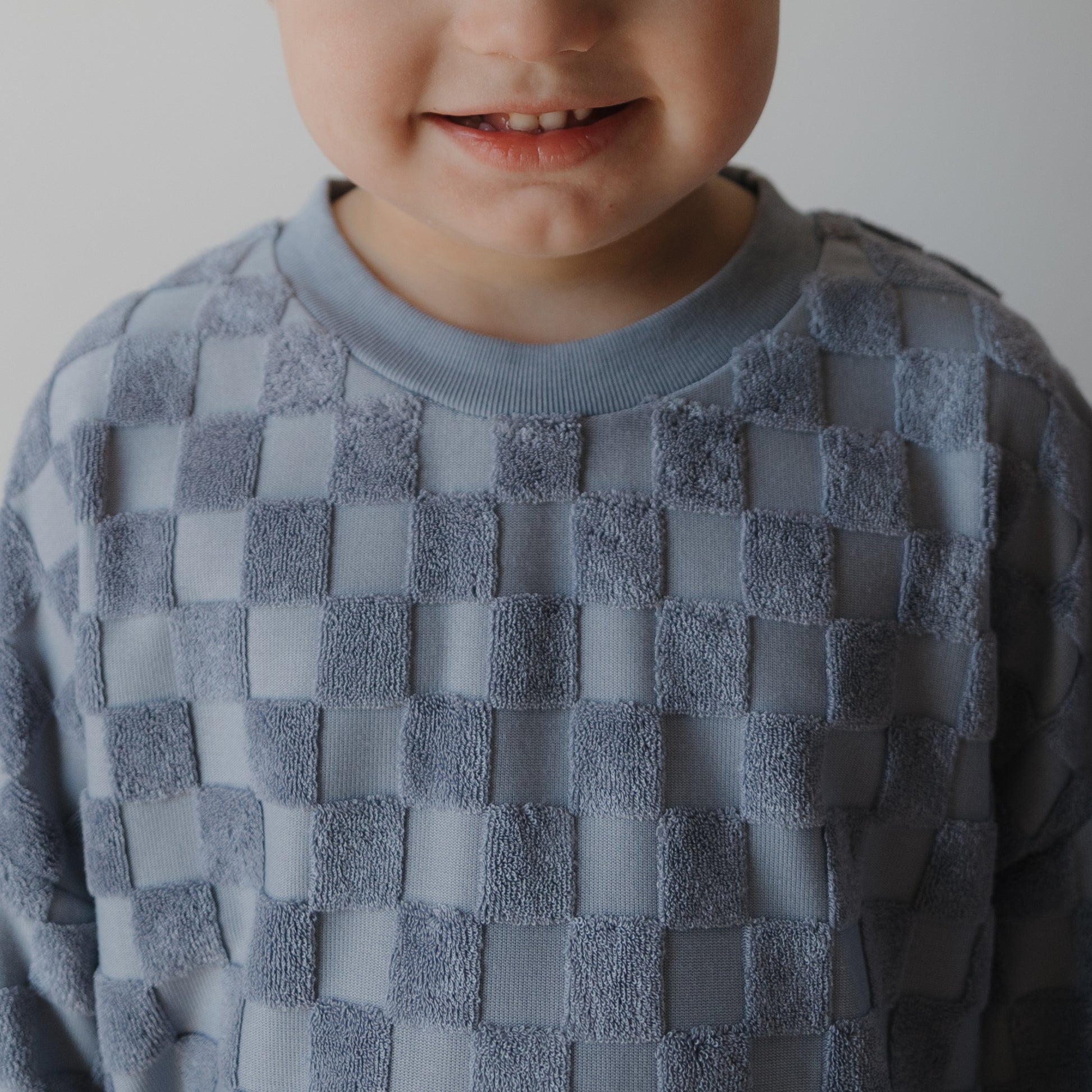 A child in the forever french baby Child Terry Short Set | Tradewinds smiles, revealing their teeth. The cropped photo shows only the lower face and upper torso against a plain background.
