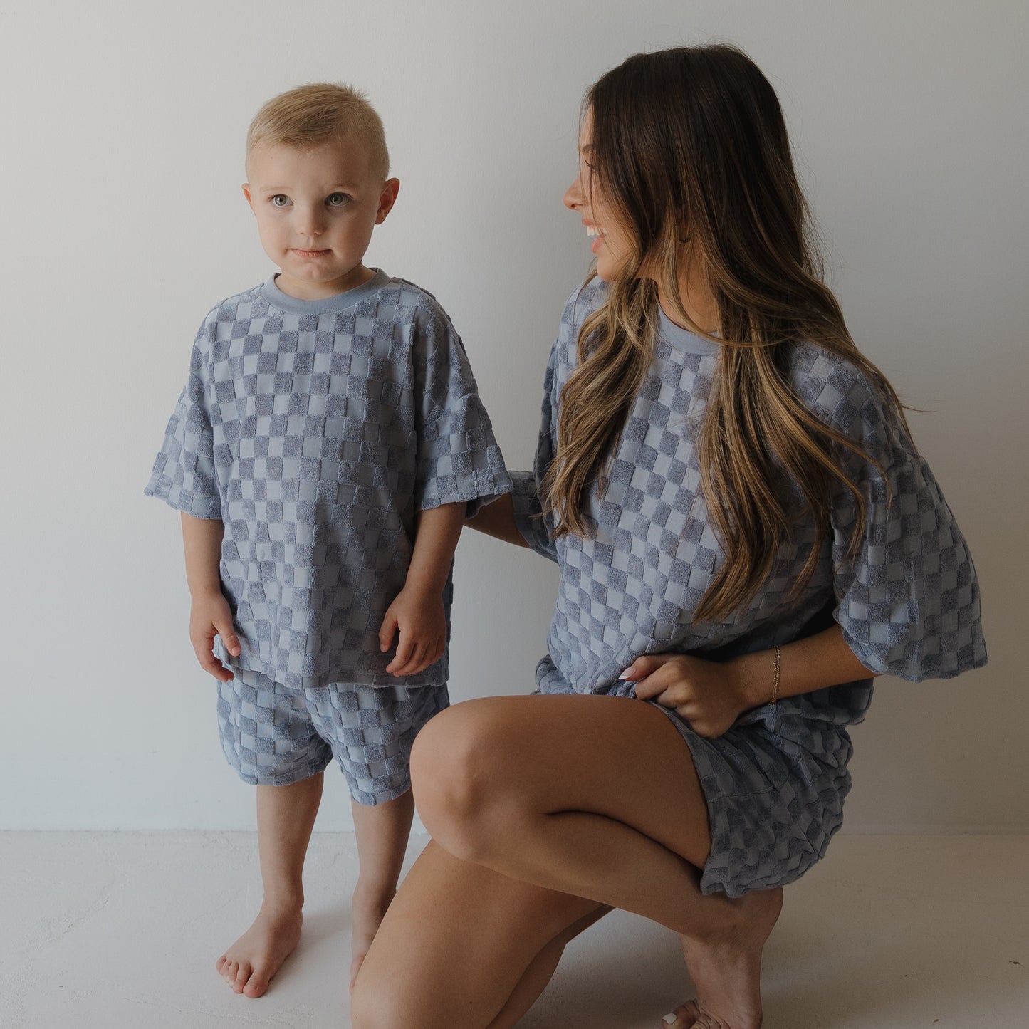 A young woman in the forever french baby Women's Terry Short Set | Tradewinds kneels barefoot beside a small boy. Both wear matching blue and gray checkered outfits as she smiles at him and he looks at the camera.