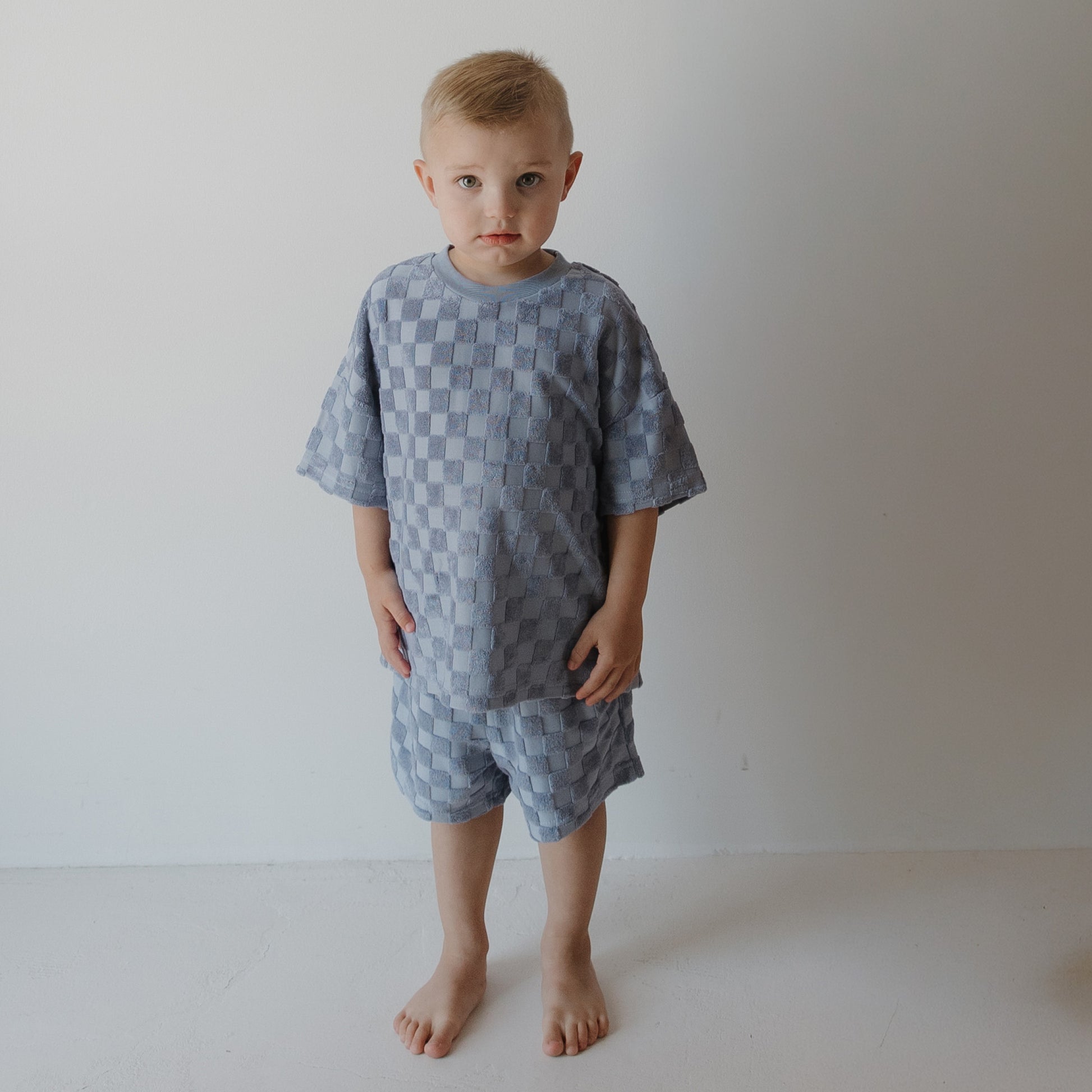 A young child with short blond hair stands barefoot indoors, wearing the forever french baby Child Terry Short Set | Tradewinds, looking slightly upward with a neutral expression against a plain white background.
