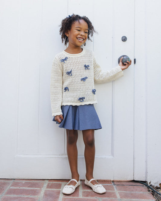 A young girl smiles as she stands in front of a white door, dressed in the Rylee + Cru Crochet Knit Pullover | Bows—a cream 100% cotton sweater adorned with blue designs. She pairs it with a blue skirt and white shoes, her short curly hair topped with a small bow.