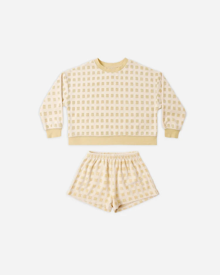 Introducing the Karli Set by Rylee + Cru: a coordinated ensemble featuring a cream-colored, long-sleeve sweatshirt and matching shorts adorned with a subtle light yellow checkered pattern. Crafted from soft terry cloth, this set offers an easygoing coastal vibe and is displayed laid flat on a white background.